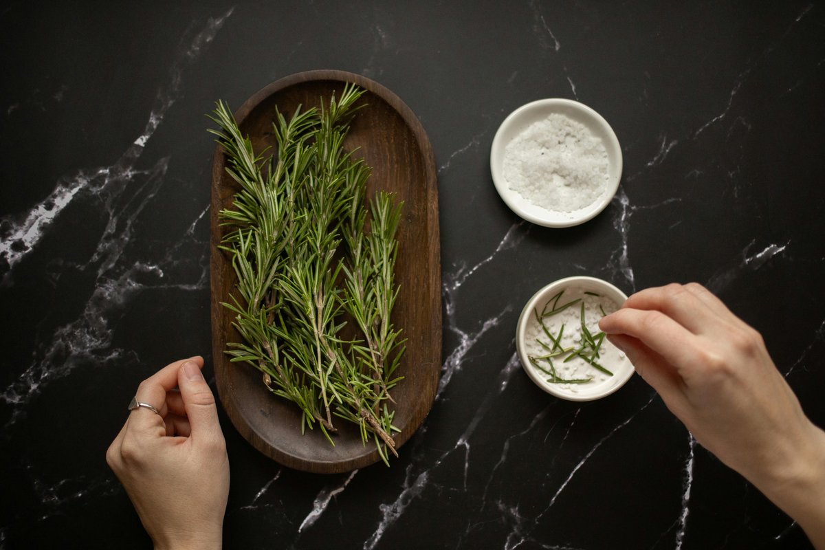 Hands arranging rosemary sprigs on a wooden tray with bowls of sea salt and olive oil on dark marble, ingredients for Greek olive oil body scrub.